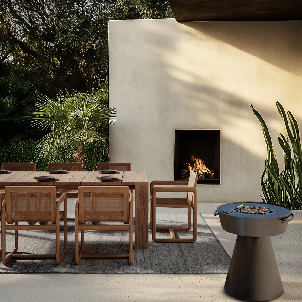 Lemieux et Cie Dock Teak Dining Armchair in teak with woven panel back, outdoor dining chair on patio beside plaster fireplace and palms