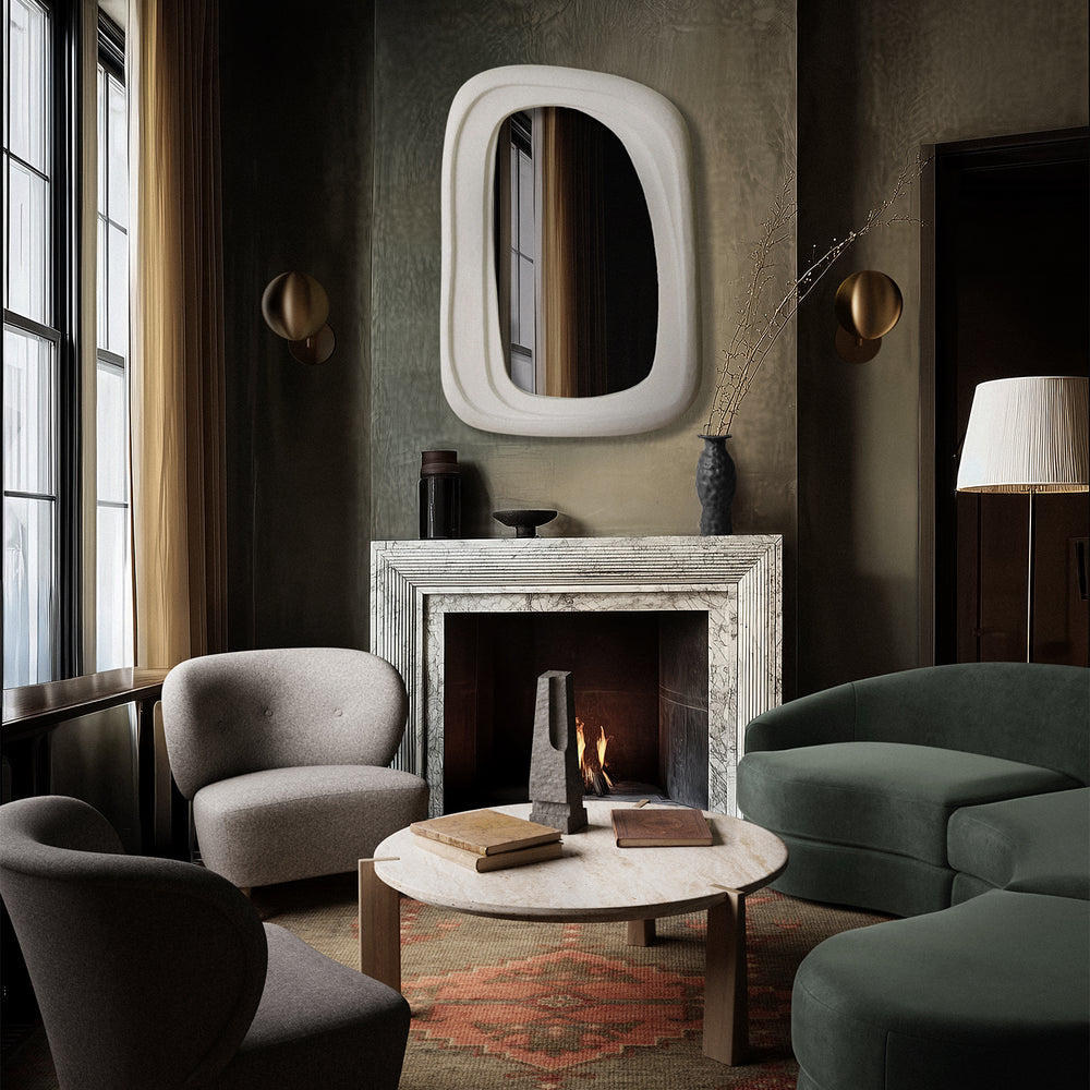 Lemieux et Cie Tourenne Chair in moss green, rounded sculptural lounge chair with tapered legs, styled beside neutral upholstered chair and round stone-top table in dark-walled sitting room with marble fireplace.