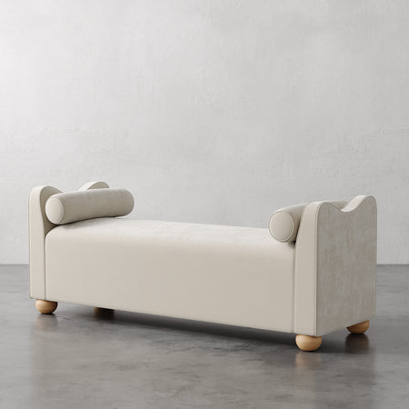 Lemieux et Cie Danielle Upholstered Bench in ivory with sculptural armrests and bolster pillows, rounded feet, shown on concrete floor against neutral plaster walls.