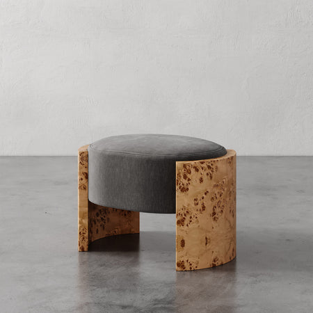 Lemieux et Cie Benoit Ottoman with charcoal upholstered seat and figured burl oak curved base, shown on polished concrete floor against textured plaster walls.