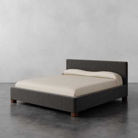 Lemieux et Cie Gérard Bed in charcoal upholstery with low-profile platform frame, shown with ivory bedding on polished concrete floor against neutral walls.