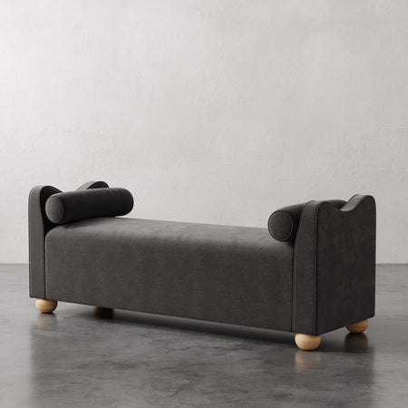 Lemieux et Cie Danielle Upholstered Bench in charcoal with cylindrical bolsters and turned legs, shown on polished concrete floor against white walls.