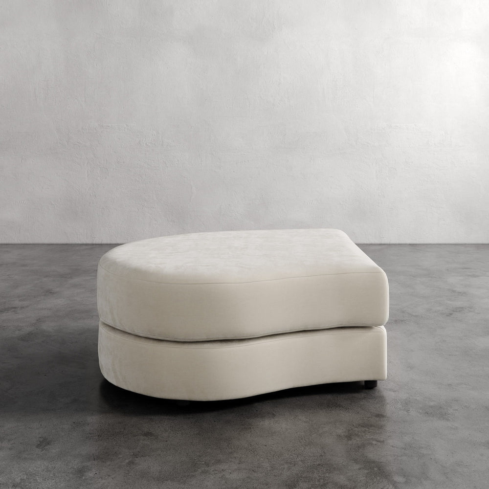 Lemieux et Cie Rulmann Left Ottoman in ivory, curved modular ottoman with rounded edges, shown on concrete floor against textured plaster walls.