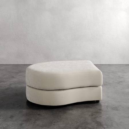 Lemieux et Cie Rulmann Left Ottoman in ivory, curved modular ottoman with rounded edges, shown on concrete floor against textured plaster walls.