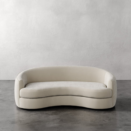 Lemieux et Cie Rulmann Sofa in ivory, curved silhouette sofa with organic sculpted form, set on polished concrete floor against textured plaster wall.