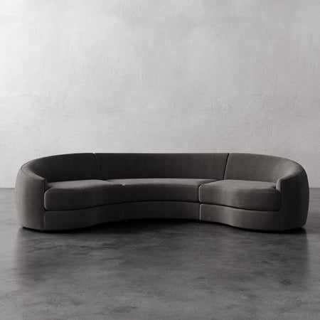 Lemieux et Cie Rulmann 3-Piece Sectional, curved sectional sofa in charcoal with rounded arms and low profile, on polished concrete floor against textured plaster walls.