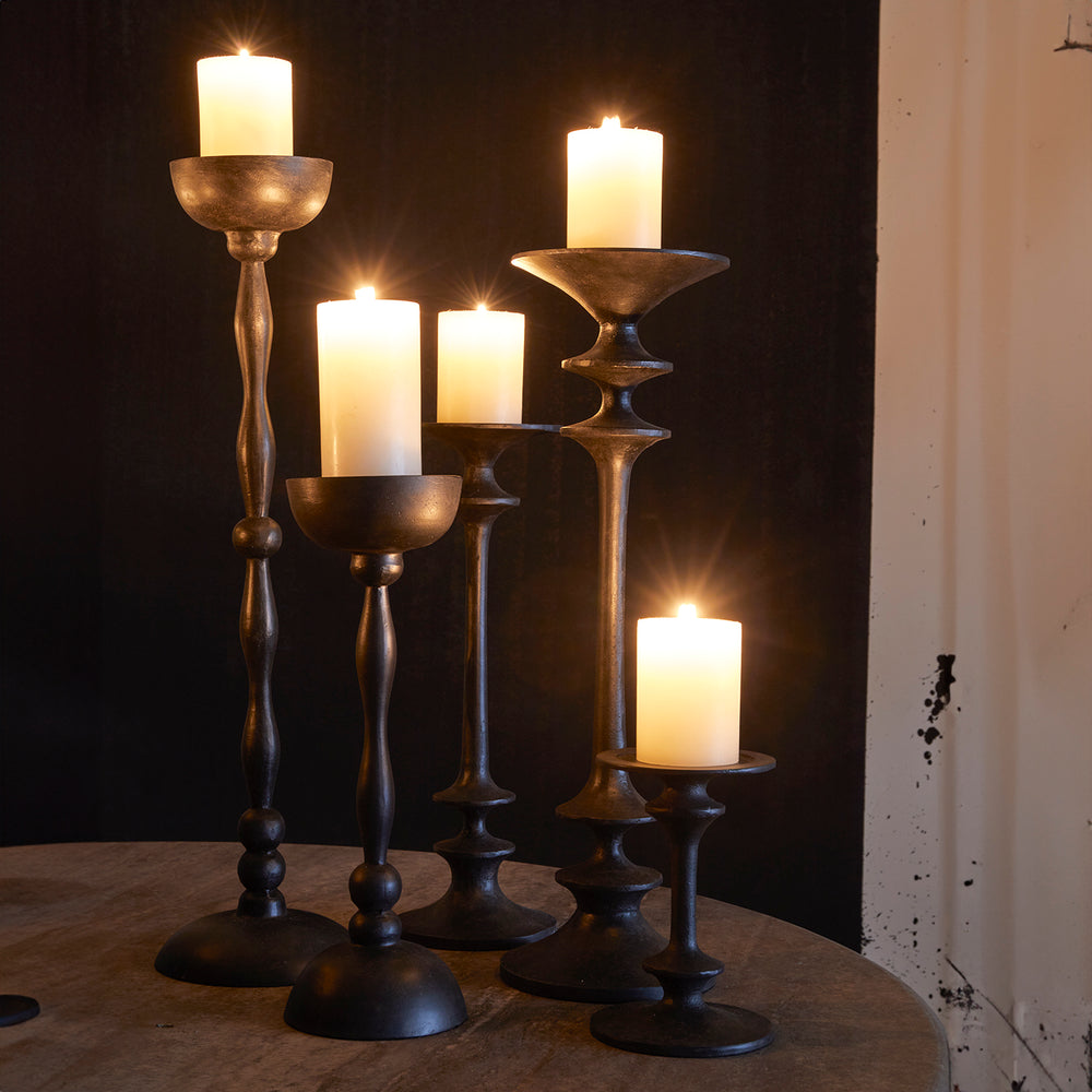 Lemieux et Cie Théophile Candle Holders in dark hand-forged finish, sculptural candlesticks with turned forms, arranged on wood table against textured plaster wall.