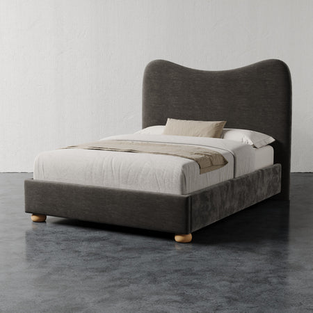 Lemieux et Cie Danielle Bed with sculptural curved headboard and rounded legs, upholstered bed dressed in neutral linens, shown on concrete floor against white walls.
