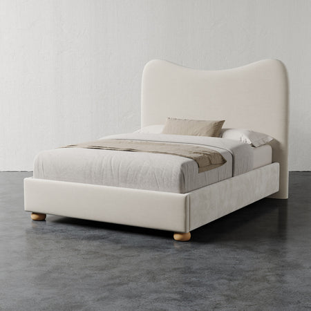 Lemieux et Cie Danielle Bed in ivory, upholstered bed with sculptural curved headboard and turned feet, shown on dark concrete floor against white plaster walls.