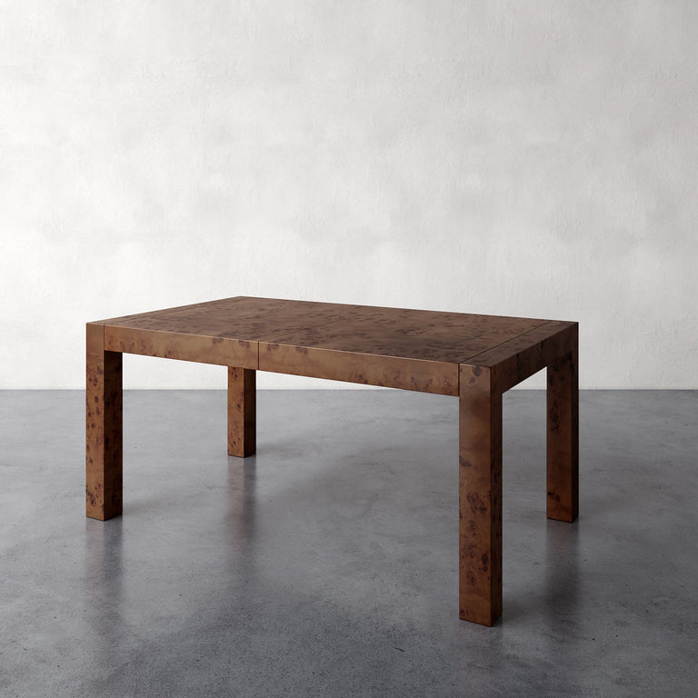 Lemieux et Cie Avenir Extension Dining Table in warm burl wood with Parsons silhouette, shown on concrete floor against neutral plaster walls.