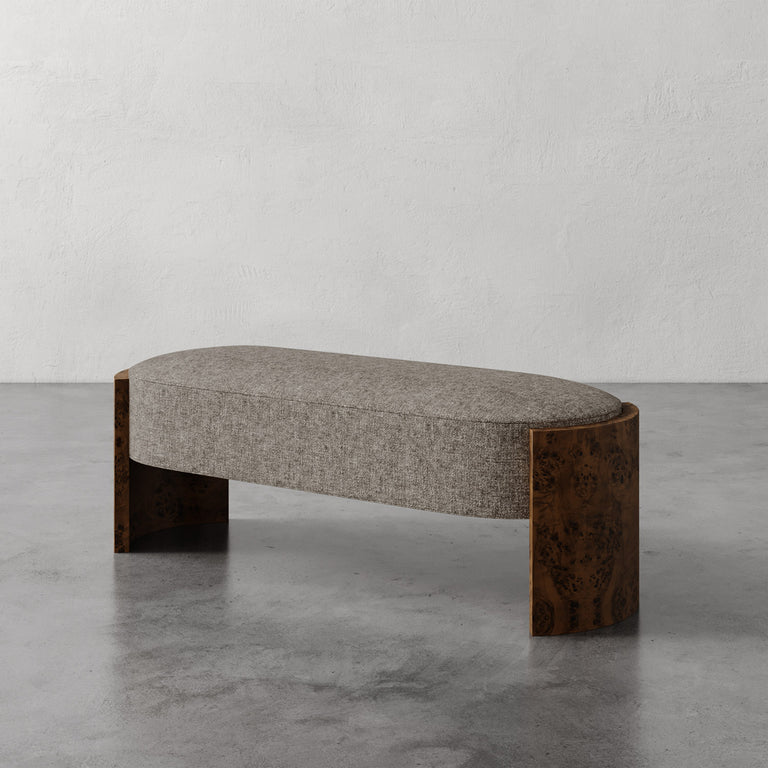 Lemieux et Cie Benoit Custom Upholstered Bench with curved maple burl legs, gray upholstered seat, shown on concrete floor against plaster walls.