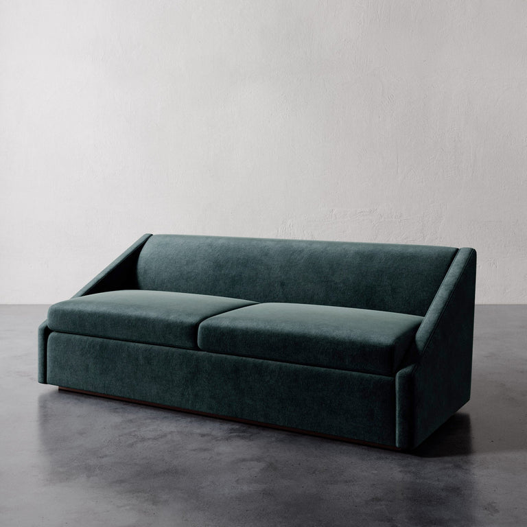 Lemieux et Cie Fontaine Sofa in deep teal upholstery, architectural sofa with angled arms, shown on polished concrete floor against white plaster wall.