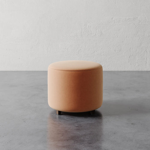 Lemieux et Cie Fabien 22" Round Ottoman in warm neutral upholstery, cylindrical form on dark base, shown on concrete floor against textured plaster wall.
