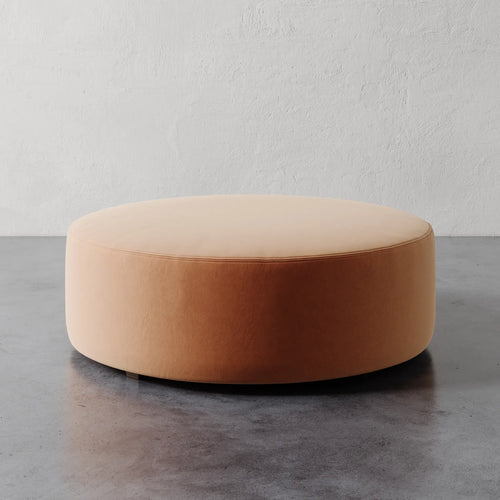 Lemieux et Cie Fabien 48" Round Ottoman in warm blush upholstery, low-profile circular ottoman on textured concrete floor against white plaster walls.
