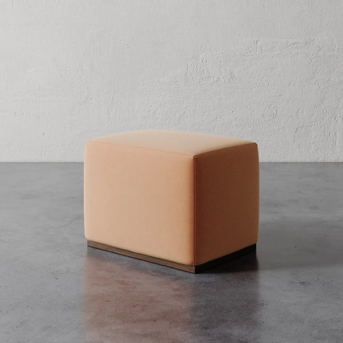 Lemieux et Cie Sophie Rectangular Ottoman in warm leather upholstery, low-profile cube ottoman on dark plinth base, shown on polished concrete floor against textured plaster wall.