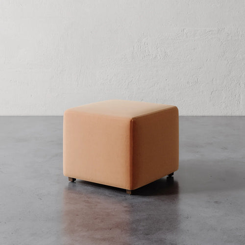 Lemieux et Cie Fabien 22" Square Ottoman in warm peach upholstery, compact ottoman on dark concrete floor against textured white plaster wall.