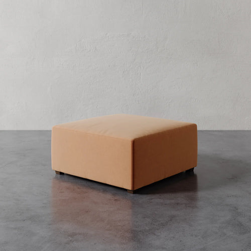Lemieux et Cie Fabien 36" Square Ottoman in warm leather, low-profile upholstered ottoman on stone floor against textured plaster wall.