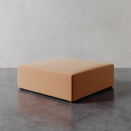 Lemieux et Cie Fabien 48" Square Ottoman in warm tan, low-profile square ottoman on concrete floor against textured plaster wall.