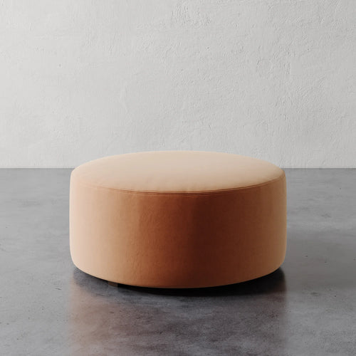 Lemieux et Cie Fabien 36" Round Ottoman, low-profile upholstered ottoman in warm peach tones, shown on concrete floor against textured plaster wall.