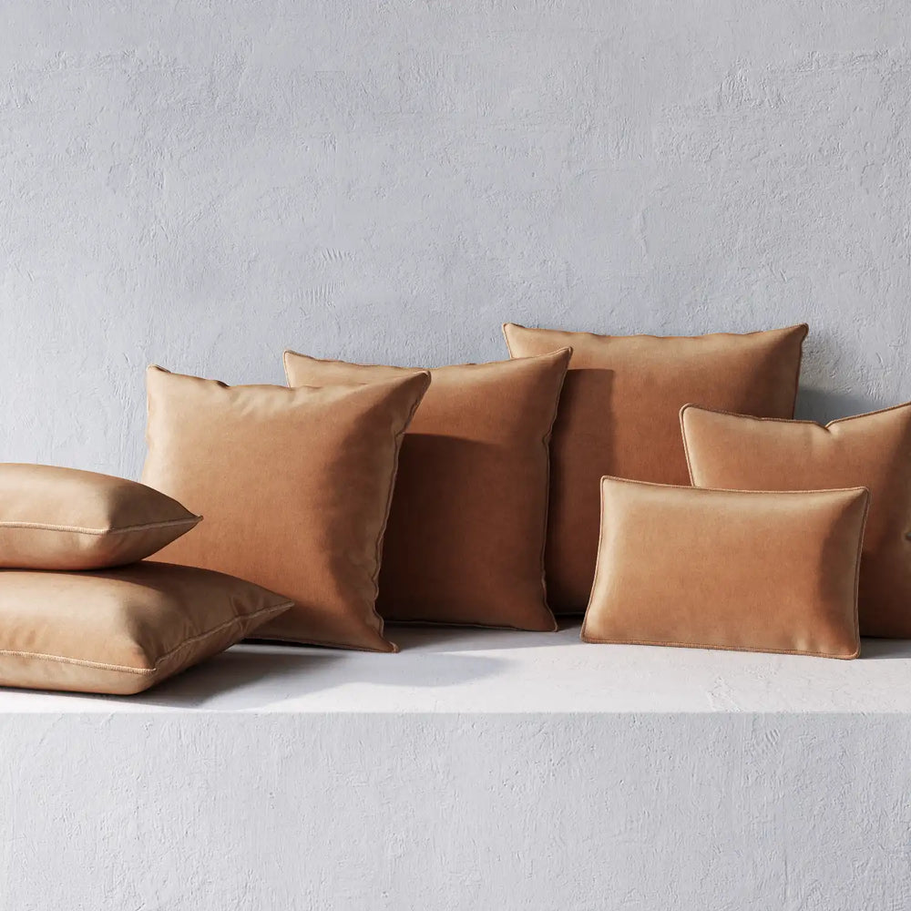 Lemieux et Cie Throw Pillows in warm tan leather, welted cushions in varied sizes, arranged against textured plaster wall on white surface.