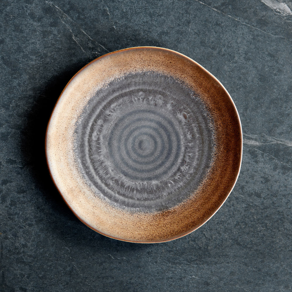 Lemieux et Cie Akita Stoneware Salad Plate in warm copper and charcoal reactive glaze, ceramic plate with concentric pattern, on dark stone surface.