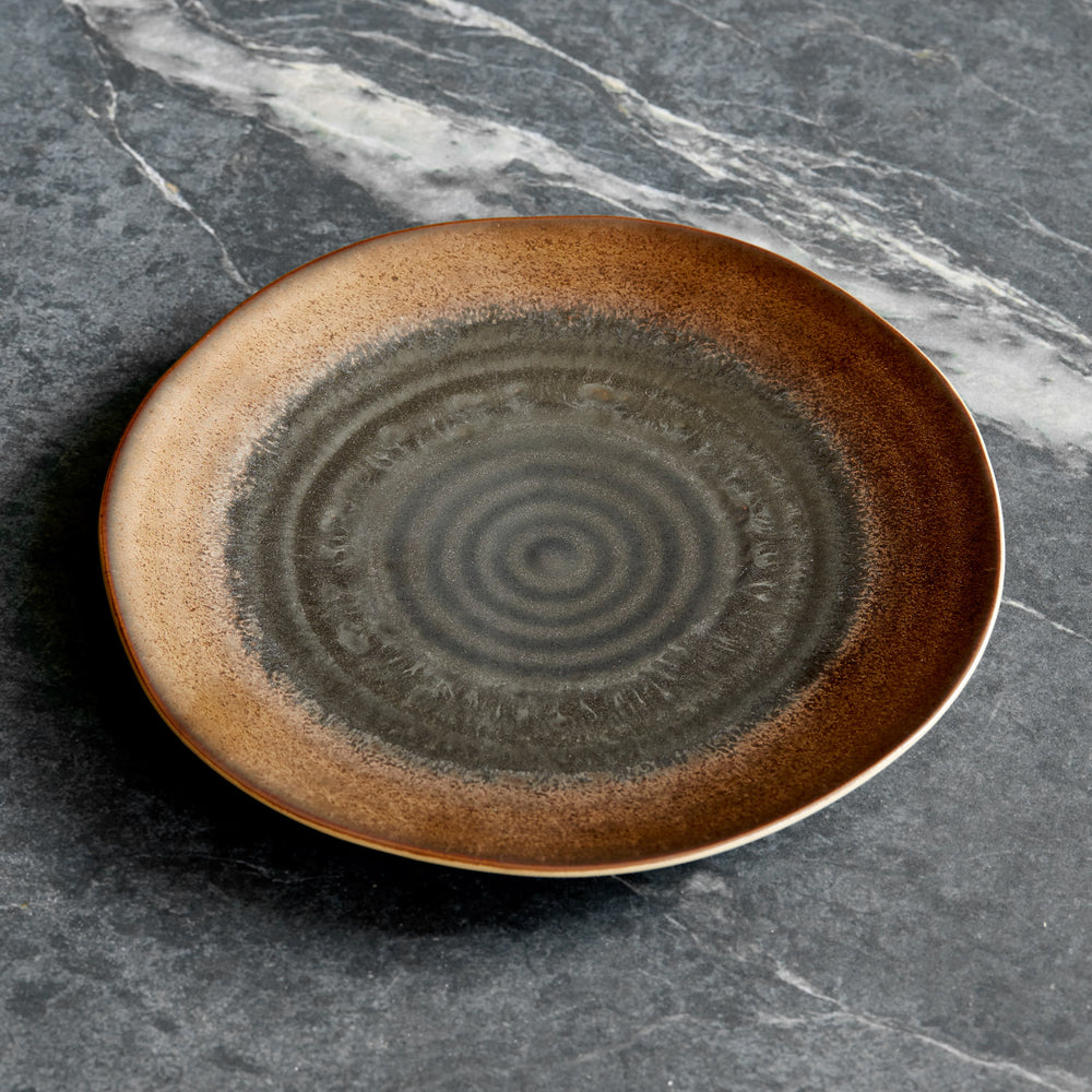Lemieux et Cie Akita Stoneware Salad Plate with reactive glaze in copper and charcoal tones, ceramic dinnerware shown on grey veined marble surface.