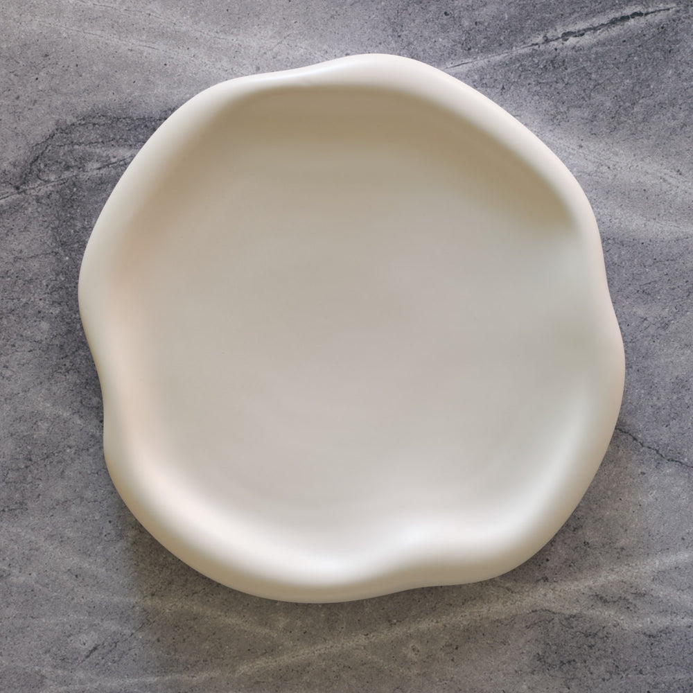 Lemieux et Cie Amorphous Dinner Plate in soft white matte stoneware, organic curved form shown on textured concrete surface.