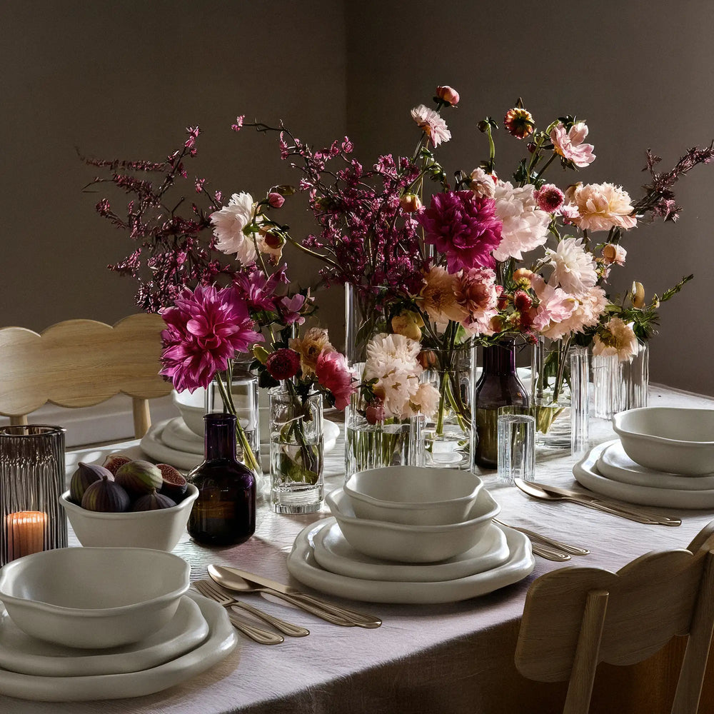 Lemieux et Cie Amorphous Dinnerware Set in soft white matte stoneware, organic-edged plates styled on linen-draped dining table with glass vases and warm daylight.