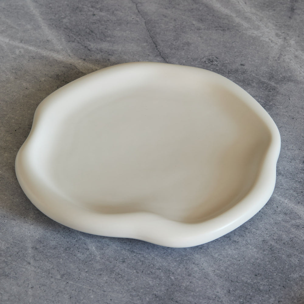 Lemieux et Cie Amorphous Dinner Plate in soft white matte stoneware, organic curved edge, shown on gray concrete surface.