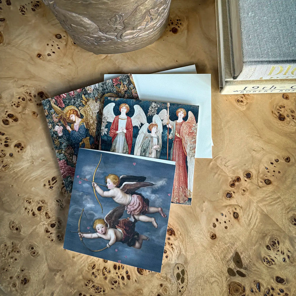 Lemieux et Cie Angels Jubilee Greeting Card Set, stacked notecards with celestial imagery on burl wood surface beside glass bowl and fabric swatches in warm light.