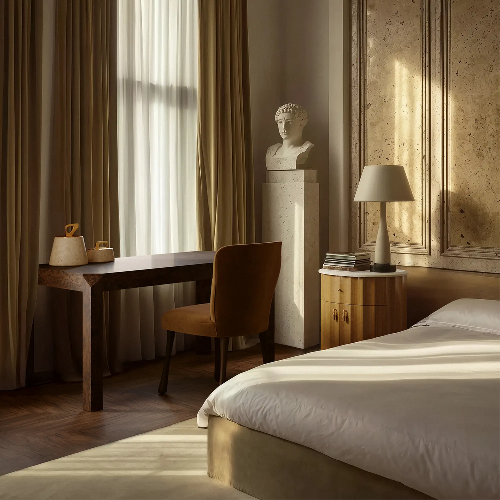 Lemieux et Cie Armand Desk styled in a warm bedroom with tan drapery, stone walls, soft daylight, and a sculptural table lamp on wood floors.