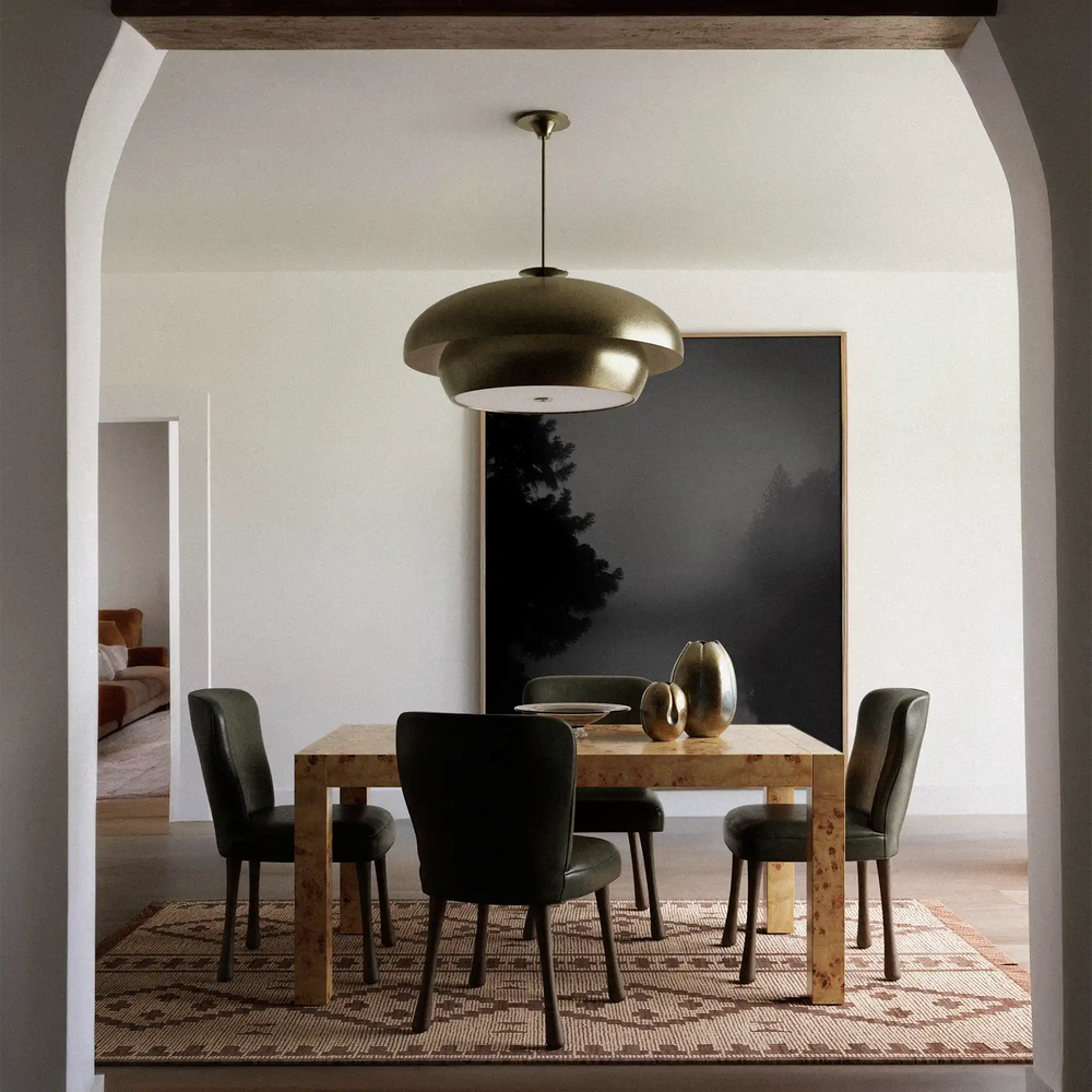 Lemieux et Cie Solene Chandelier in hand-gilded brass, sculptural rounded pendant hung above dining table with upholstered chairs, white arched walls, framed landscape art.