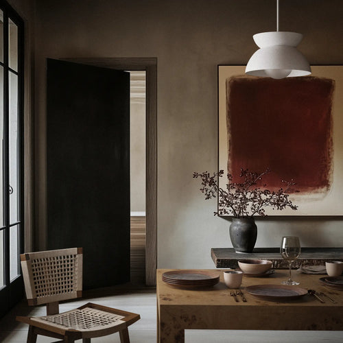 Lemieux et Cie Jeanneret Dining Chair in teak with hand-woven cane seat, shown in warm-lit dining room with plaster walls and abstract art.