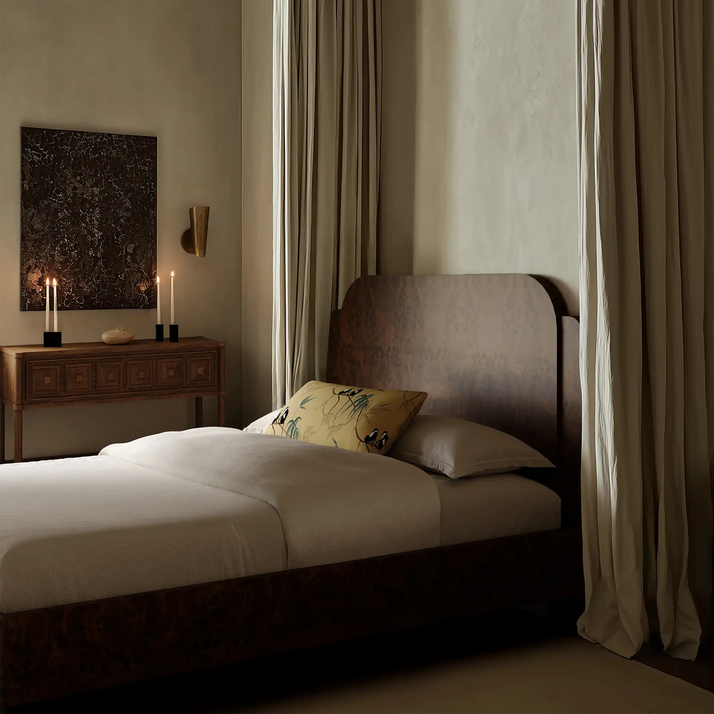 Lemieux et Cie Avenir Bed with curved headboard and neutral linen bedding, styled in a plaster-walled bedroom with soft drapery and warm candlelight.