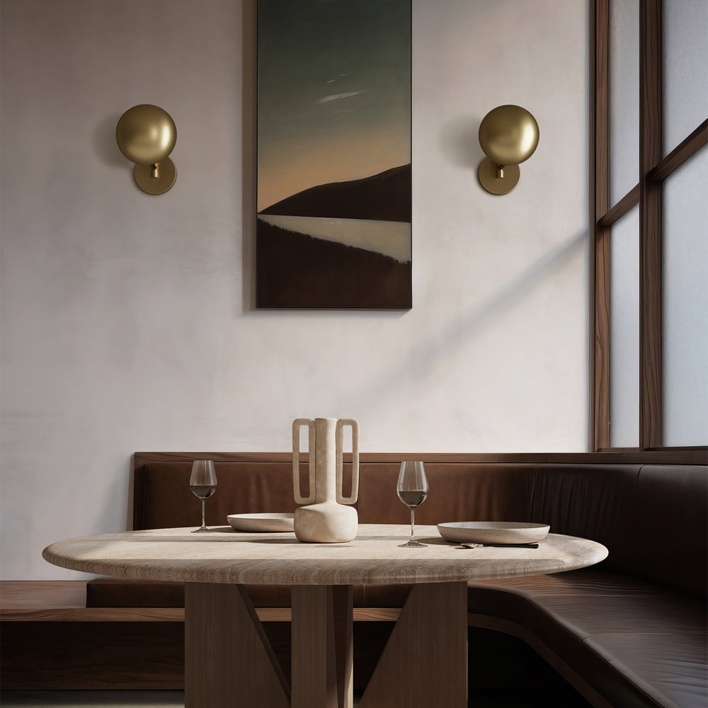 Lemieux et Cie Balleroy Medium Sconce , a luxury lighting fixture lighting a cozy dining area with the Elysées Dining Table, curved seating, wall art, and warm brass details.