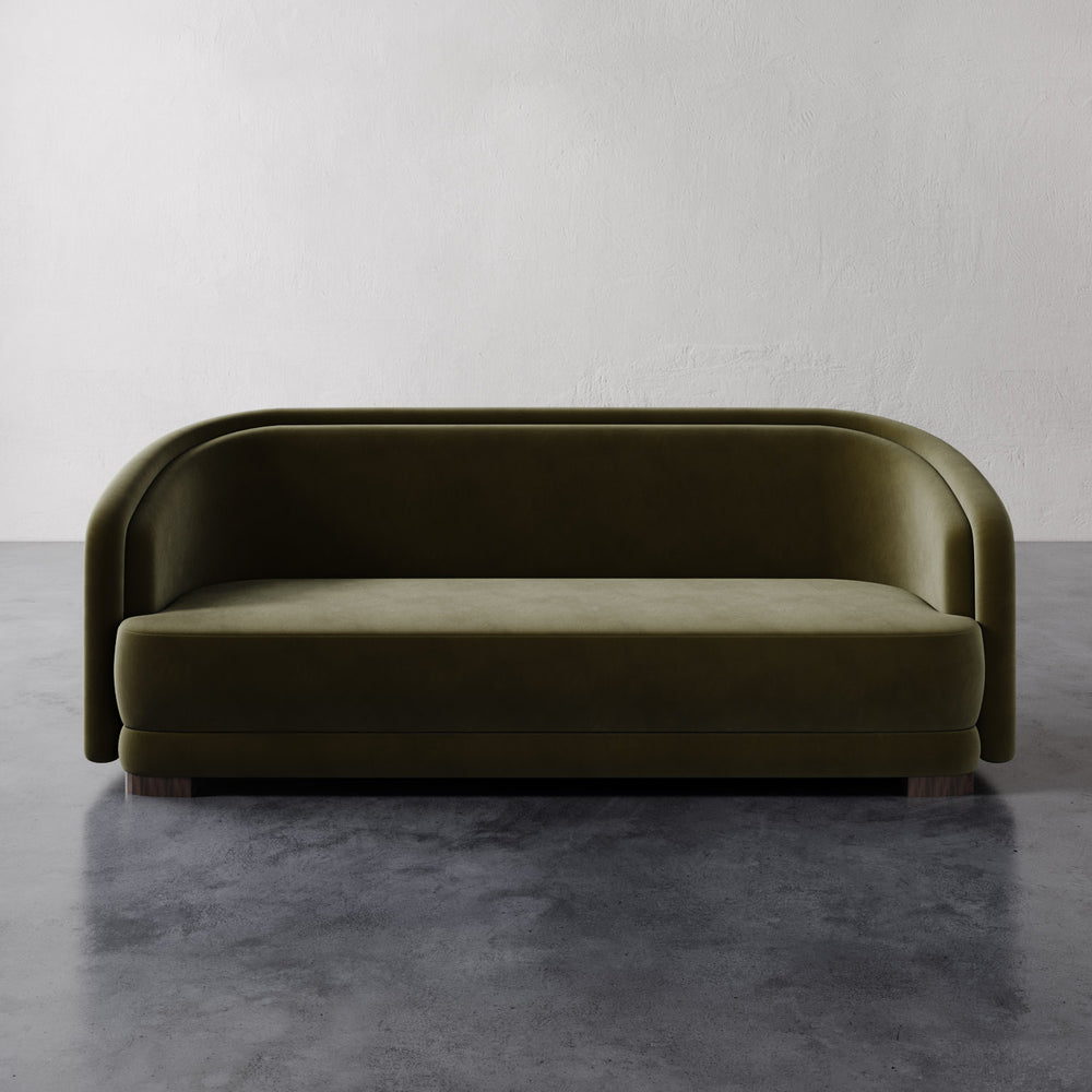 Lemieux et Cie Valence Sofa in olive green, low-profile sofa with rounded arms and plinth base, shown on dark concrete floor against white walls.