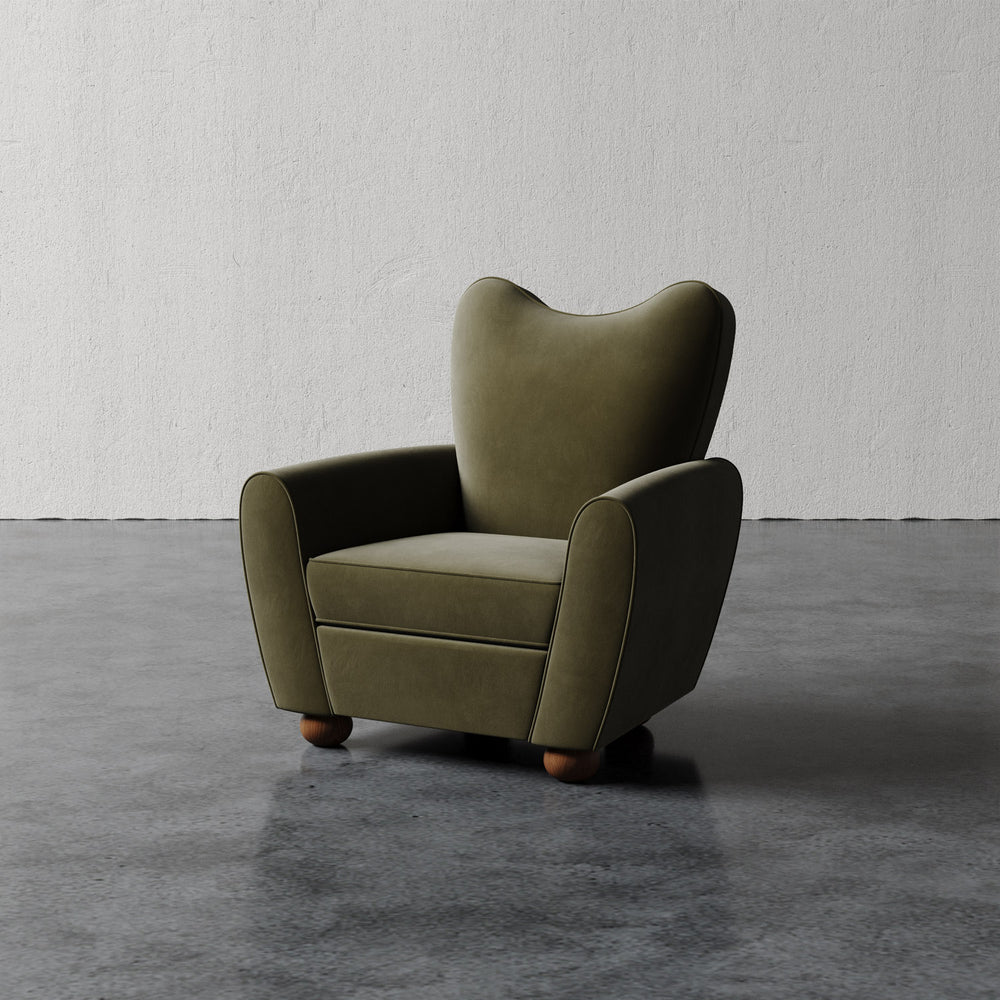 Lemieux et Cie Danielle Chair in olive green, sculptural wingback form with rounded arms and turned legs, shown on polished concrete floor against textured plaster wall.