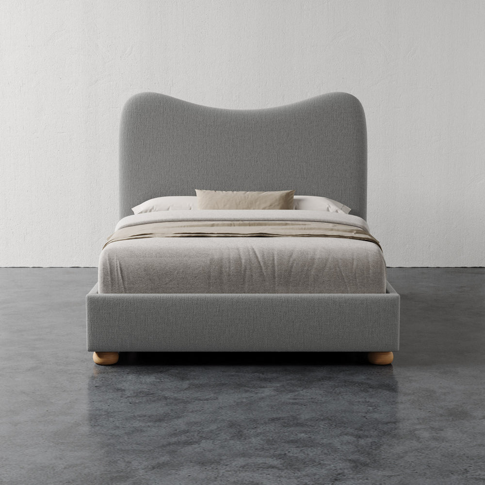 Lemieux et Cie Danielle Bed with curved sculptural headboard in grey, upholstered bed styled with neutral linens on dark concrete floor.