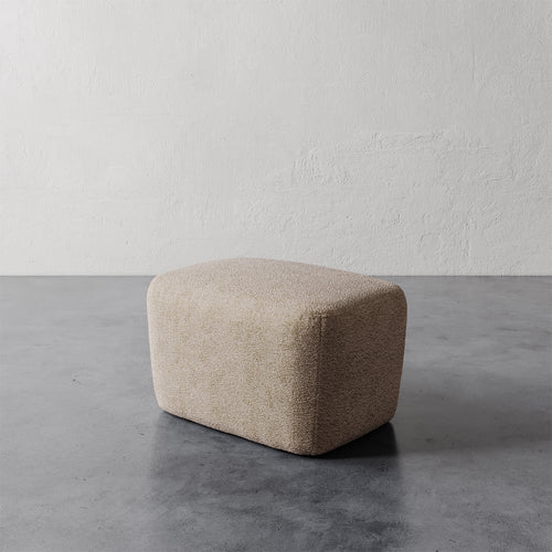 Lemieux et Cie Aurélie Ottoman in warm beige, low-profile rounded ottoman on concrete floor against textured plaster walls.
