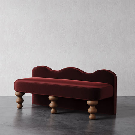 Lemieux et Cie Celine Bench in deep burgundy upholstery with sculptural wave back and turned sphere legs, shown on dark concrete floor against white plaster wall.