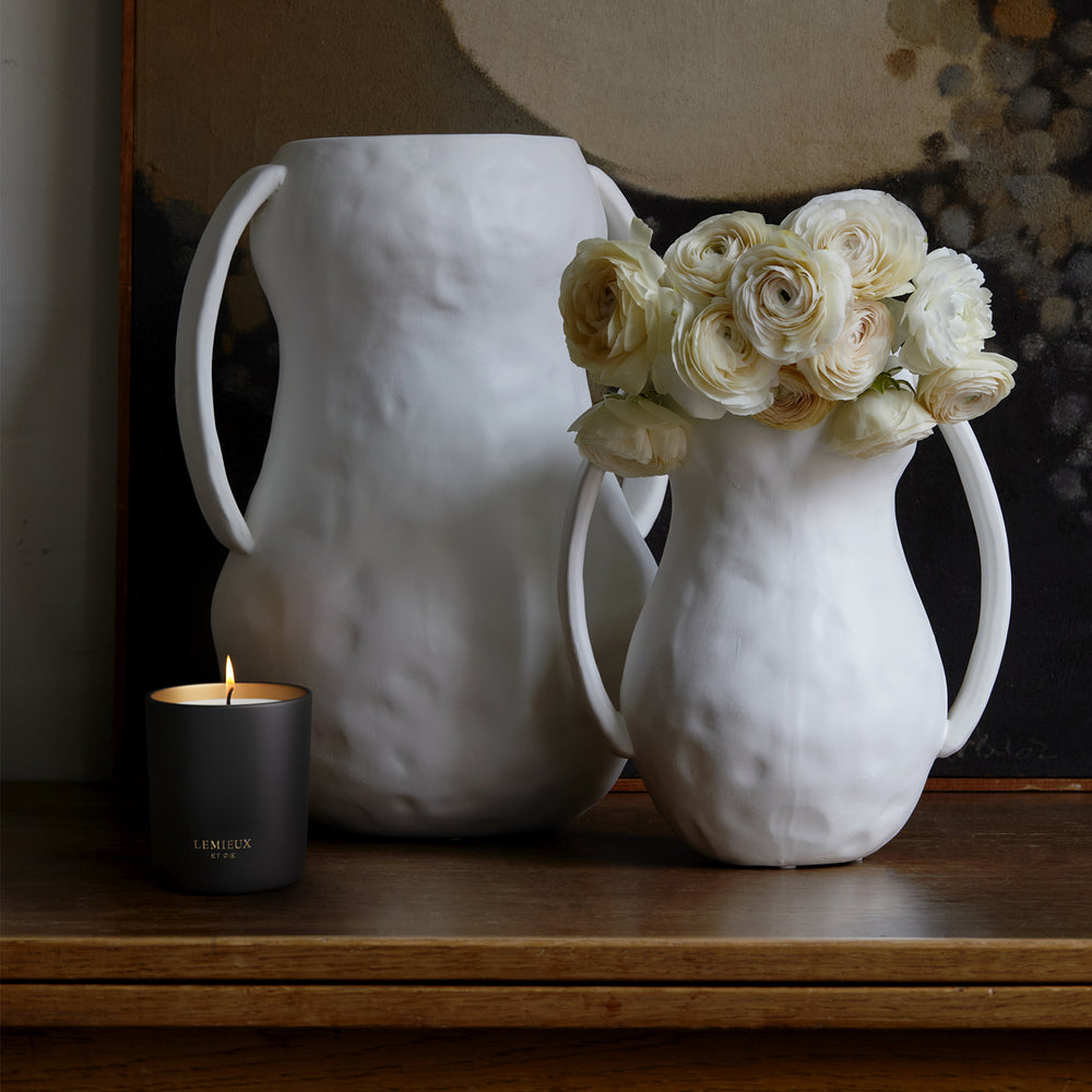 Lemieux et Cie La Côte d'Acadie Filled Candle in matte dark vessel with ivory ranunculus, styled on wood surface with abstract art.