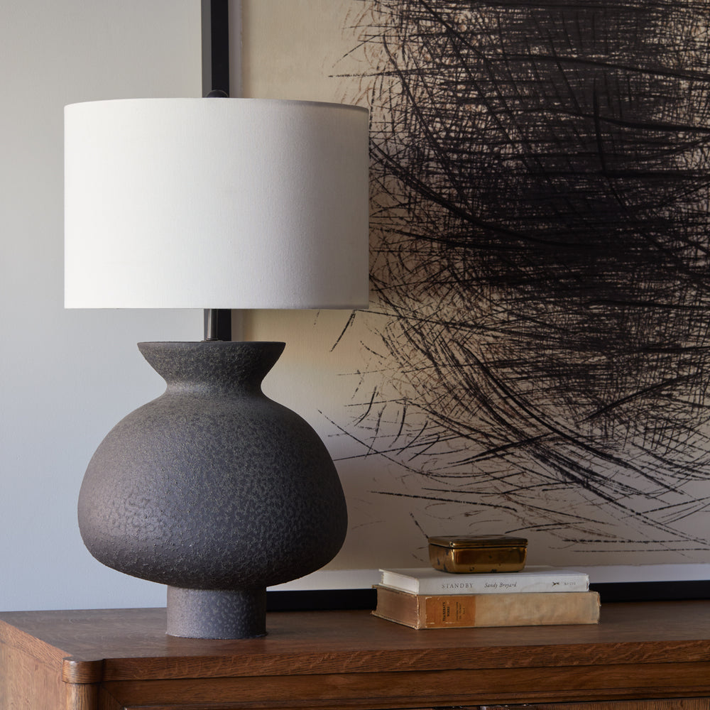 Lemieux et Cie Casis Lamp in matte black, sculptural earthenware table lamp with rounded shade, styled on wood surface with artwork and books.