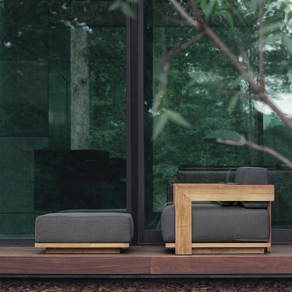 Lemieux et Cie S2 Teak Ottoman with charcoal cushions and solid teak frame, outdoor seating on a wood deck beside glass-walled pavilion surrounded by greenery.