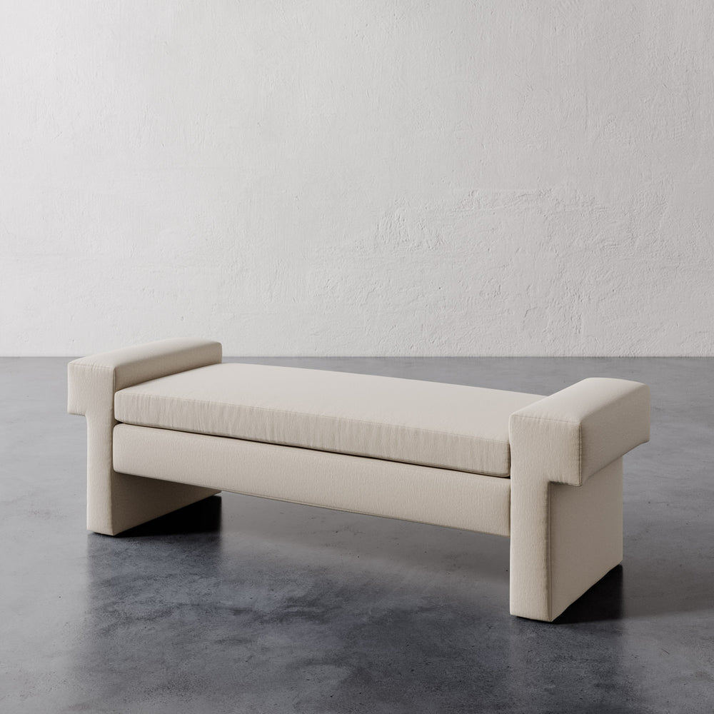 Lemieux et Cie Lemaire Bench in ivory, upholstered bench with blocky geometric arms, shown on dark concrete floor against textured plaster wall.