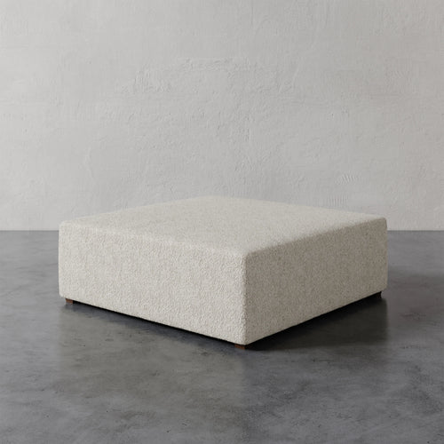 Lemieux et Cie Fabien 48" Square Ottoman in ivory bouclé, low-profile seating piece on polished concrete floor against textured plaster walls.