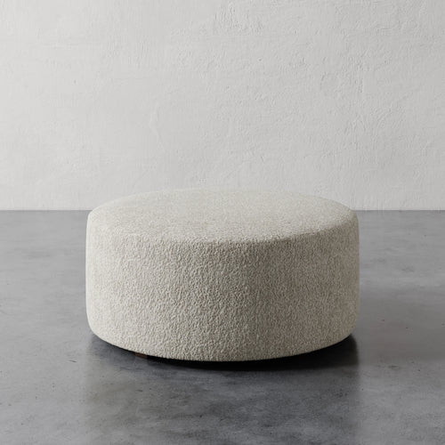Lemieux et Cie Fabien 36" Round Ottoman in ivory bouclé, low-profile cylindrical ottoman on concrete floor against textured plaster walls.