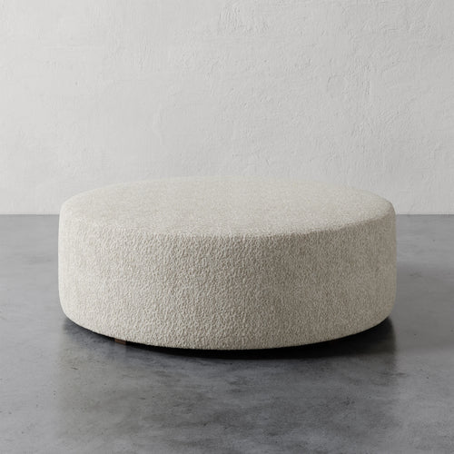 Lemieux et Cie Fabien 48" Round Ottoman in ivory bouclé, low-profile circular ottoman on concrete floor against textured plaster walls.