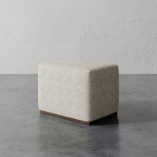 Lemieux et Cie Sophie Rectangular Ottoman in ivory bouclé, low-profile ottoman on dark base, shown on concrete floor against textured plaster walls.
