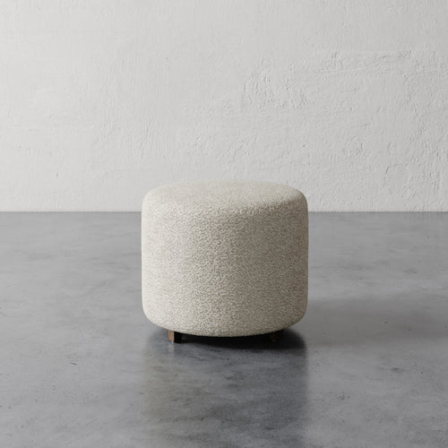 Lemieux et Cie Fabien 22" Round Ottoman in ivory bouclé, low cylindrical ottoman on polished concrete floor against textured plaster walls.