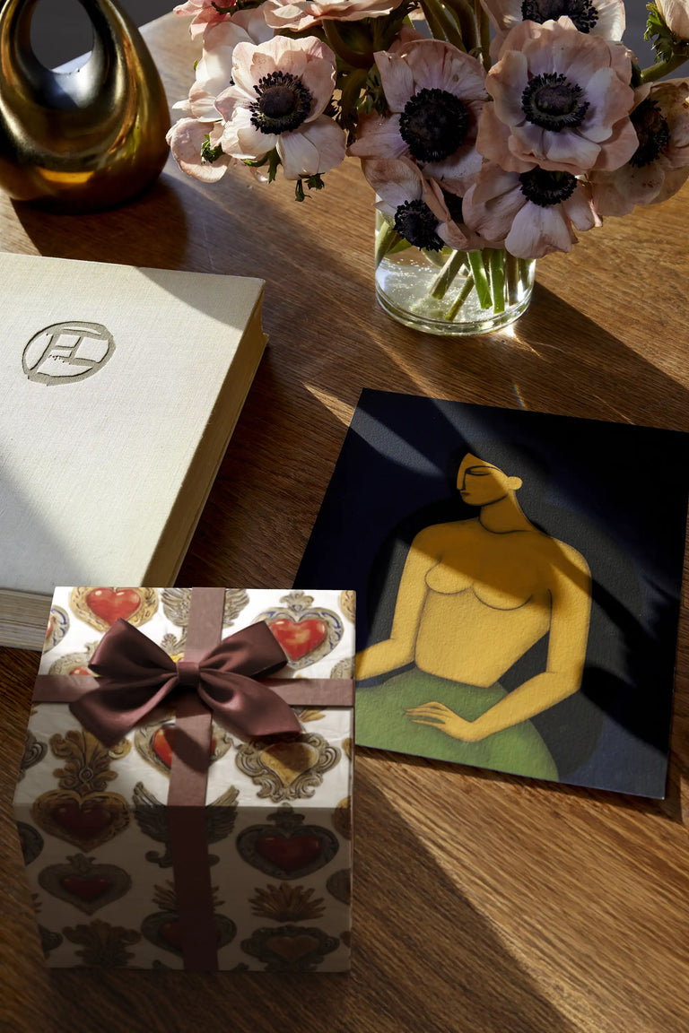 Lemieux et Cie Gift box wrapped in Corazón Sagrado luxury gift wrapping paper inspired by Mexican folk art, next to a vase of flowers, and Les Femmes artwork on a wooden surface